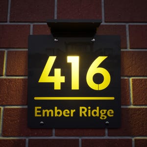May include: Black rectangular address sign with the number "416" in yellow, and the words "Ember Ridge" below. The sign is mounted on a red brick wall and has a black solar panel above it.