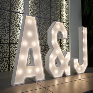 May include: White marquee letters spelling out "A & J" with illuminated bulbs. The letters are standing on a tiled surface in front of a decorative patterned wall. The letters are large and create a warm, inviting glow.