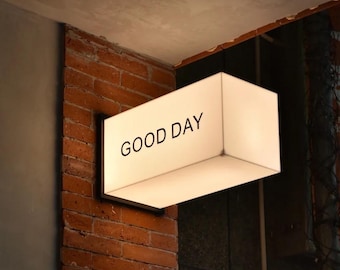 May include: A rectangular, illuminated sign with the words "GOOD DAY" in black lettering. The sign is mounted on a brick wall. The sign is white and glows, suggesting it is lit from within.