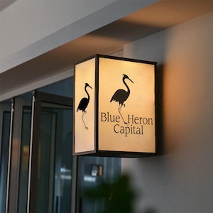May include: A rectangular, illuminated sign with the text "Blue Heron Capital" and a silhouette of a heron. The sign is black with a cream-colored background and is mounted on a wall.