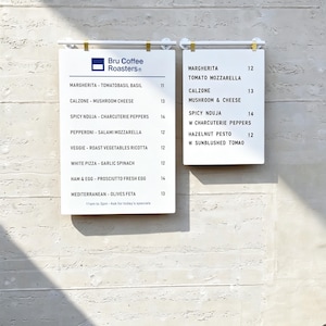 May include: Two white rectangular menu boards with black text mounted on a wall. The larger board displays the Bru Coffee Roasters logo and a list of pizza options with prices. The smaller board lists additional food items and their prices.