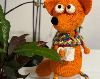 Puede incluir: Juguete de zorro de crochet naranja sosteniendo una taza blanca. El zorro tiene ojos grandes blancos y una nariz negra. El zorro lleva una bufanda de crochet colorida.