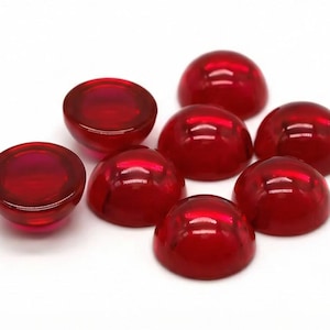 May include: A collection of eight translucent, cabochon-cut, ruby-red gemstones. The polished stones are dome-shaped with a flat base, reflecting light. These gemstones are likely used for jewelry making or other crafts.