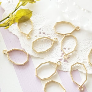 Op de afbeelding: Een groep van tien gouden metalen achtkantige hangerframes met een lus aan de bovenkant. De frames zijn gerangschikt op een witte kantonderlegger en een gestreept oppervlak.