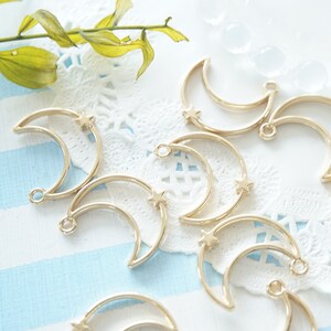 May include: A collection of gold-toned metal crescent moon charms with a small star detail. The charms are arranged on a white lace doily and a blue and white striped surface.