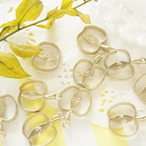 May include: A collection of ten gold-toned metal apple-shaped pendant frames. The frames are open and have a smooth, shiny finish. They are arranged on a white lace doily with a yellow and white striped background.