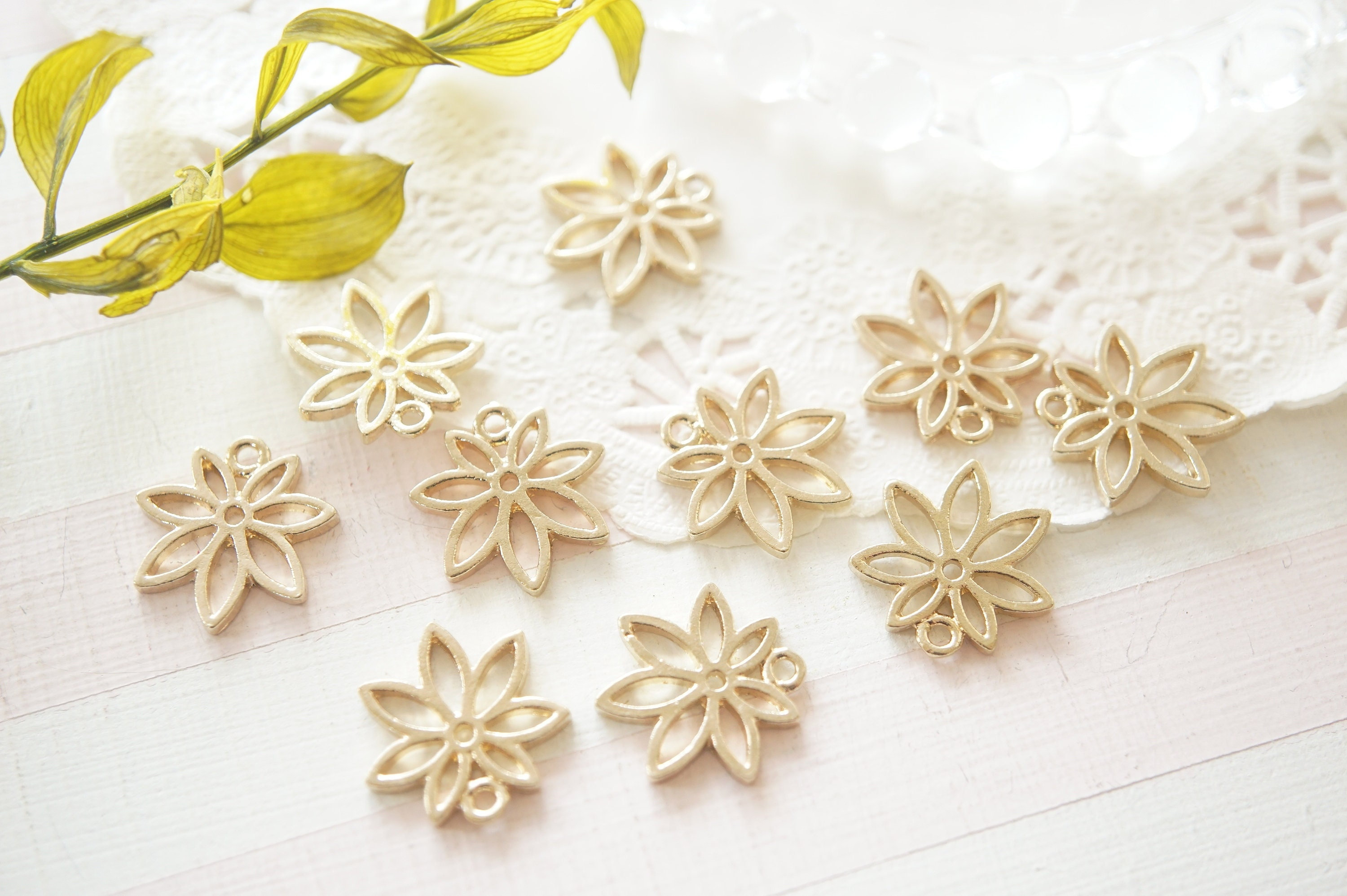 May include: Ten gold metal flower charms with a delicate openwork design. The charms are arranged on a white lace background.