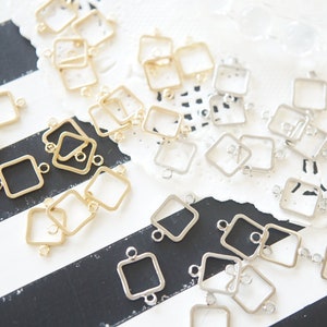 May include: A collection of gold and silver square-shaped metal frames with two jump rings on each frame. The frames are arranged on a black and white striped surface.