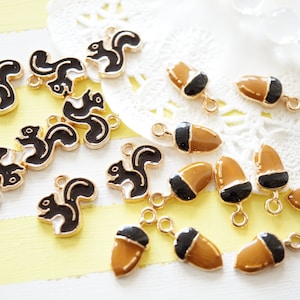 Peut inclure: Collection de breloques en métal doré représentant des écureuils noirs et blancs et des glands bruns et noirs. Les breloques ont de petites boucles pour les attacher à des bijoux ou autres créations. Le fond est un mélange de dentelle blanche et de rayures jaunes.