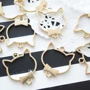May include: A set of gold metal cat-shaped charms with a bow detail. The charms are on a black and white striped surface.