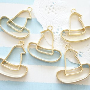 May include: Five gold-coloured metal witch hat shaped charms with clear resin inserts. The charms are on a white and blue striped background.