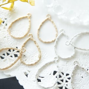 May include: Close-up of several teardrop-shaped metal jewellery findings in gold and silver. The findings have a textured hammered finish and small loops for attaching to other components. They are arranged on a white lace doily.
