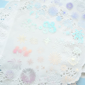 May include: A sheet of iridescent white paper doilies with a variety of firework designs printed on them. The doilies are layered on top of each other and have a delicate, lacy pattern.