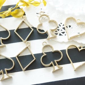 Puede incluir: Encantos de metal dorado en forma de corazones, diamantes y palos de cartas de juego. Los encantos están sobre una superficie rayada en blanco y negro.