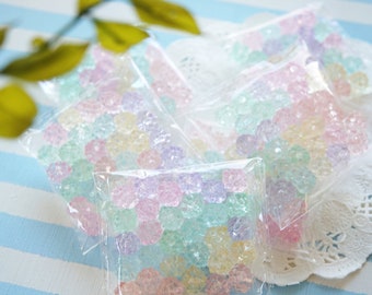 May include: Multiple small bags filled with pastel-colored, translucent, flower-shaped beads. The bags are arranged on a white doily and a blue and white striped surface.