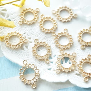 May include: Gold-colored metal sunburst charms with a central open circle and decorative edges. These charms are arranged on a white lace doily with a blue and white striped background. The charms have small loops for attaching to jewelry.