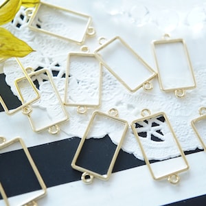 May include: A group of gold-toned metal rectangular frames with two jump rings on each frame. The frames are scattered on a white lace doily and a black and white striped surface.