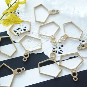May include: A set of ten gold-toned metal hexagon-shaped frames with a small loop at the top and a decorative ring at the bottom. The frames are arranged on a white lace doily and a black and white striped surface.