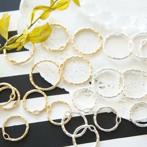 Op de afbeelding: Een verzameling gouden en zilveren springringen, met een getextureerde afwerking. De ringen zijn gerangschikt op een wit kantdoily, met een groene plantenstengel op de achtergrond.