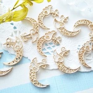 May include: Gold-coloured crescent moon charms with detailed filigree patterns and cloud-like accents. These charms have small loops for attaching to jewellery or other crafts. The charms are arranged on a white lace doily.