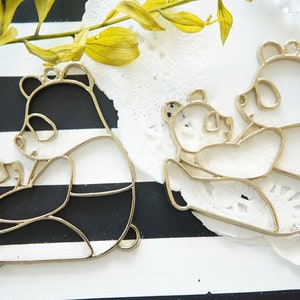 May include: Two gold metal panda bear outlines, one holding a smaller panda bear outline. The outlines are on a black and white striped background.