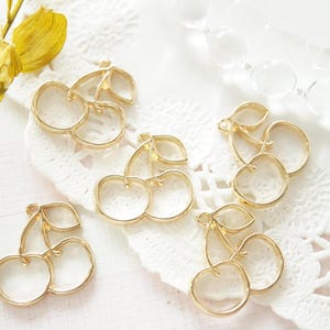 Puede incluir: Seis abalorios de cereza en metal dorado. Cada abalorio es una cereza doble con un tallo de hoja. Los abalorios están sobre un fondo blanco.