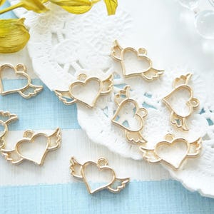 May include: Gold-colored heart-shaped charms with angel wings, arranged on a white lace doily and a light blue striped surface. The charms have a small loop at the top for attaching to jewelry or other crafts.