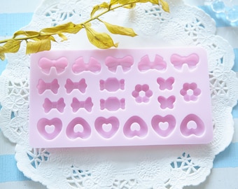 May include: A pink silicone mould featuring bow, sweet, flower, butterfly, and heart shapes. The mould is for crafting and sits on a white lace doily. The background is a blue and white striped pattern.