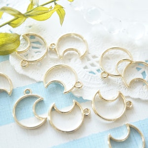 May include: A collection of gold-toned metal crescent moon-shaped pendant blanks with jump rings. The blanks are arranged on a white and blue striped surface.