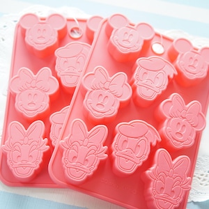 May include: Two rectangular pink silicone moulds featuring embossed character faces including Mickey Mouse, Minnie Mouse, Daisy Duck, and Donald Duck. Each mould has a hanging hole at the top and multiple cavities for shaping food or craft items.