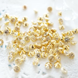 Op de afbeelding: Een stapel gouden metalen versieringen met een verhoogd midden en een decoratieve rand. De versieringen zijn verspreid over een wit kant oppervlak.