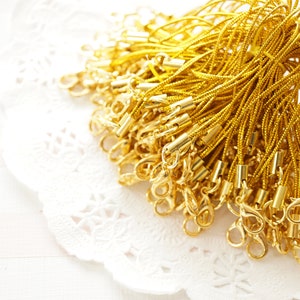 Puede incluir: Un montón de cordón dorado y cierres metálicos. Los cordones están agrupados y los cierres están unidos a los extremos de los cordones.