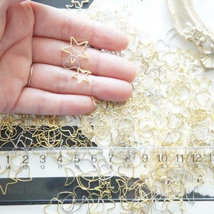 Peut inclure: Charmes dorés en métal en forme d'étoiles et autres formes, dispersés sur une surface blanche. Une règle indique les mesures en centimètres. Les breloques sont délicates et semblent faites à la main, adaptées à l'artisanat ou à la bijouterie.