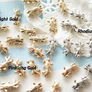 May include: Assortment of gold and silver goldfish charms. The charms are in three colors: light gold, pinkish gold, and rhodium.  The charms are scattered on a white surface with blue stripes.