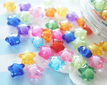 May include: A collection of colorful, star-shaped beads in various translucent hues, including blue, pink, yellow, orange, green, and purple. Some beads are scattered on a white surface, while others are in a clear glass container.