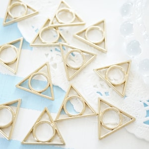 May include: A collection of gold-toned metal triangle charms with a circle cut out in the centre. The charms are scattered on a white doily and a blue and white striped surface.