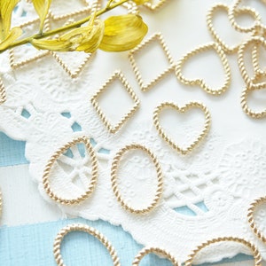 May include: Gold-colored metal charms in various shapes, including hearts, ovals, and diamonds, with a twisted rope design. The charms are scattered on a white lace doily and a light blue and white striped surface. A sprig of yellow leaves is also present.