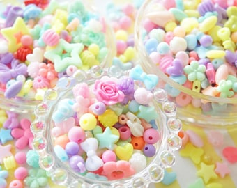 May include: A collection of colorful plastic beads in a variety of shapes, including stars, flowers, hearts, and bows. The beads are in a clear glass bowl and are perfect for crafting.