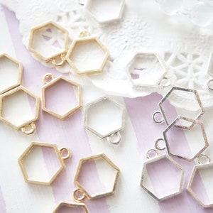May include: A collection of gold and silver hexagonal metal frames with a loop at the top. The frames are arranged on a striped purple and white surface.