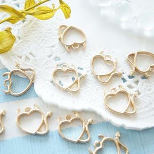 May include: A collection of gold-toned metal heart-shaped charms with small devil horns and tails. The charms are arranged on a white lace doily.