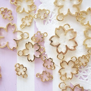 May include: A collection of gold-tone metal charms shaped like cherry blossoms. The charms are translucent and have a pink or white interior.