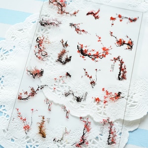 Può includere: Foglio di adesivi trasparente con illustrazioni di rami di fiori di ciliegio, con fiori rossi e rami neri. Gli adesivi sono disposti su uno sfondo bianco, con un centrino di pizzo e una superficie a righe blu e bianche.