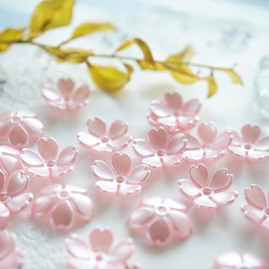 Op de afbeelding: Een verzameling delicate, lichtroze kersenbloesem bloemversieringen. De plastic bloemen zijn verspreid over een wit oppervlak, met een paar geelgroene bladeren op de achtergrond. De bloesems hebben een parelmoerglans.