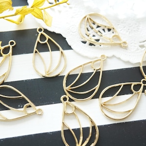 May include: Gold-colored teardrop-shaped metal charms with an openwork design. The charms have a loop at the top for attaching to jewelry. The charms are arranged on a black and white striped surface.