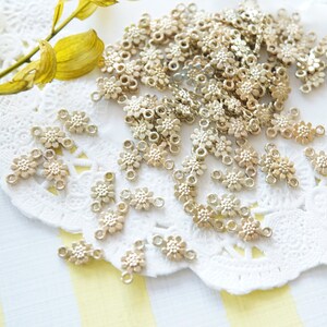Peut inclure: Un grand nombre de breloques en forme de fleurs en métal doré avec deux boucles sur chaque breloque. Les breloques sont éparpillées sur une dentelle blanche.