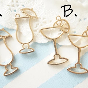 Könnte beinhalten: Zwei goldfarbene Metall-Anhänger in Form von Cocktailgläsern. Ein Anhänger zeigt ein Cocktailglas mit einem Regenschirm oben, beschriftet mit "A.". Der andere Anhänger zeigt ein Cocktailglas mit einer Zitronenscheibe am Rand, beschriftet mit "B."