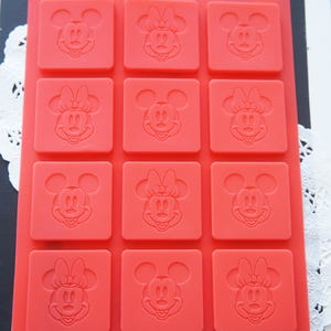 May include: A rectangular red silicone mold featuring twelve square cavities, each embossed with a character face design. The grid contains alternating patterns of Mickey Mouse and Minnie Mouse faces. The text Disney echo is printed at the bottom.