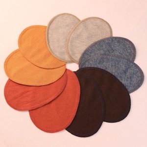 Może przedstawiać: Kolekcja dziesięciu okrągłych, materiałowych wkładek laktacyjnych w różnych kolorach, w tym pomarańczowym, rdzawym, szarym, niebieskim i brązowym. Każda wkładka ma obszytą krawędź. Wkładki ułożone są w okrąg na jasnoróżowym tle.