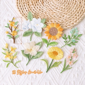 Könnte beinhalten: Eine Sammlung von sieben gestickten Blumenaufnähern mit Sonnenblumen, weißen Blüten und grünen Blättern. Ein Aufnäher ist neben einem Lineal abgebildet, das eine Höhe von 3 cm anzeigt. Der Text 23 Styles Available ist unten in Orange gedruckt.