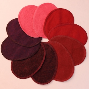 Peut inclure: Une collection de coussinets ovales en tissu de différentes nuances de rouge, rose et violet, disposés en cercle. Chaque coussinet a un bord cousu, et les couleurs varient du rose clair au bordeaux foncé.